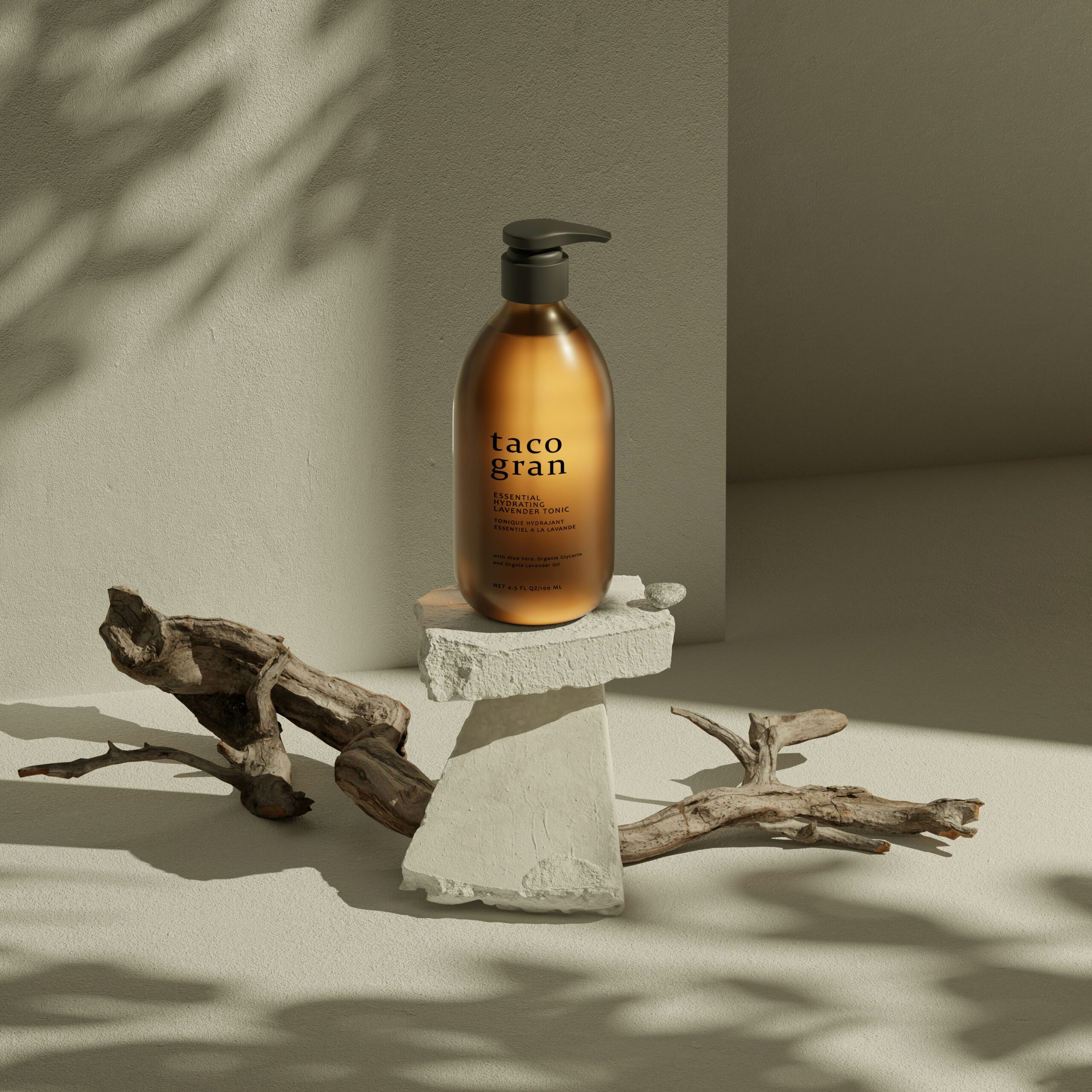 A studio shot of a brown translucent pump bottle labeled "taco gran" resting on a white stone pedestal. The scene is minimalist, featuring dry wooden branches on a neutral sandy-colored floor with soft leaf shadows cast against a beige wall.